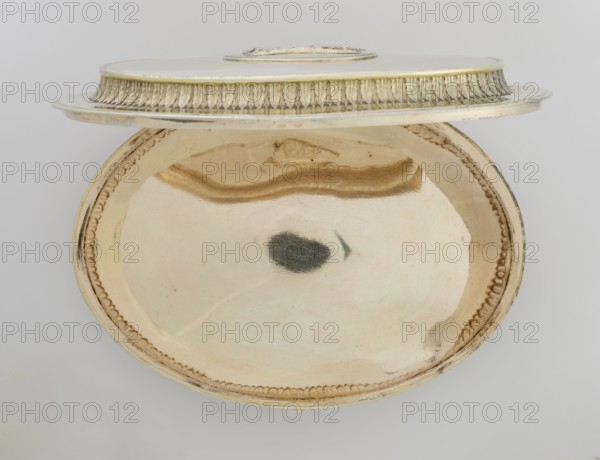 Oval toilet box by Thomas I Pitts fl. 1755–ca.1793, goldsmith. Oval box on four volute feet formed of acanthus leaves; lower edge with a band of bay leaves, above them laurel festoons with suspended medallions containing rosettes, upper edge with an anthemion frieze. Hinged recessed cover bordered with a band of bay leaves matching the body; central medallion on the cover with a relief of four putti at a sacrificial altar. Classicism/Georgian/George III style; relief decorations, festoons, palmettes, anthemions, bay leaves. Dated 1780–1781. England, London, Great Britain. Toilet accessory. Height 12 cm; width 27.1 cm; depth 20 cm; weight 1627 g., A round metallic dish with a lid shown partly open, the interior of the dish reflecting a smooth surface with a few darker smudges and a curved reflective streak; the lid has decorative ridged edging and a raised central area. The visible colors include gold, beige, cream, silver, and a few dark gray reflections against a pale gray background.