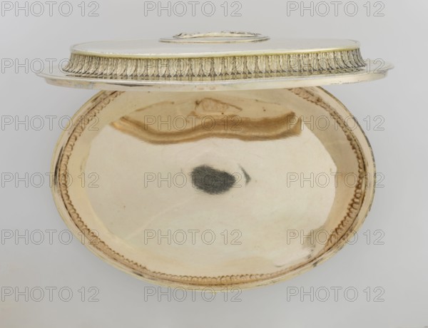 Oval toilet box by Thomas I Pitts fl. 1755–c.1793. Silver; Georgian/George III style, 1780/1781, London, England. Oval box on four volute feet formed of acanthus leaves. Lower edge decorated with a band of bay leaves; above, laurel festoons with suspended medallions containing rosettes; upper edge with an anthemion frieze. Hinged recessed cover bordered with a band of bay leaves matching the body. Central medallion on the cover with a relief of four putti at a sacrificial altar. Ornament: festoons, palmettes, anthemions, bay leaves; relief decorations. Function: toilet accessory. Dimensions: height 12 cm, width 27.1 cm, depth 20 cm; weight 1627 g., A shallow oval metal dish with a raised rim and a matching lid set slightly ajar, both showing a smooth reflective interior and decorative edging; the dish and lid are mainly pale gold and warm beige with areas of darker golden brown reflections and a small dark grayish spot near the center of the dish; the rim decoration and lid trim show a...
