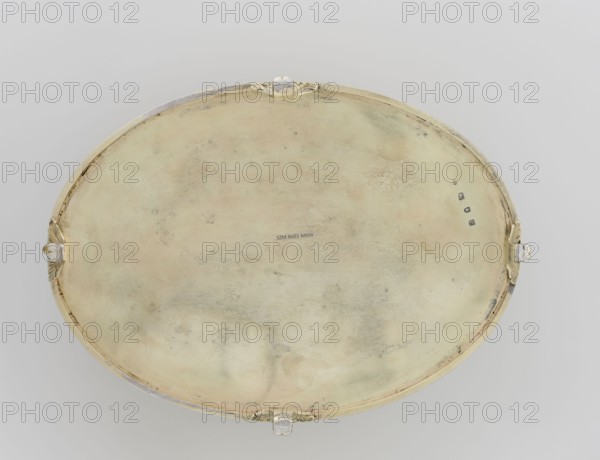 Oval toilet box by Thomas I Pitts fl. 1755–ca.1793, goldsmith; oval box on four volute feet formed of acanthus leaves. Lower edge decorated with a band of bay leaves; above them laurel festoons with suspended medallions containing rosettes; upper edge with an anthemion frieze. Hinged, recessed cover bordered with a band of bay leaves matching the body. Center of cover bears a medallion with a relief of four putti at a sacrificial altar. Classical/Georgian/George III style; relief decorations, festoons, palmettes, anthemions, bay leaves. Dated 1780–1781, London, England. Toilet accessory. Height 12 cm; width 27.1 cm; depth 20 cm; weight 1627 g., An oval metallic tray with a worn, slightly mottled surface in pale gold and muted beige tones, showing areas of darker grayish tarnish and subtle greenish discoloration; the tray has a raised rim in similar pale gold with a few decorative tab-like clasps around the perimeter that show brighter silver and gold highlights, and the background is a...