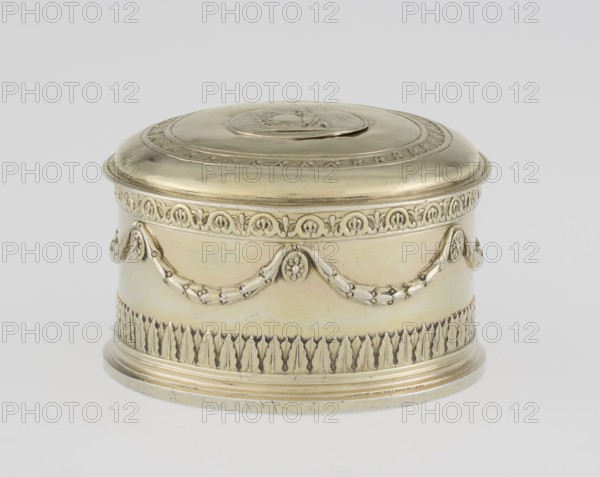 Toilet box by Thomas I Pitts fl. 1755–c.1793, goldsmith; Classicism style, Georgian/George III period, c.1780–1781, London, England. Round with lid, Classicist toilet box decorated with laurel festoons suspended from rosette medallions; lower edge with a knurled belt of stylized lesbian cymatium. Slightly arched cover edged with a knurled wavy ornament running dog, centrally bearing a circular medallion with a relief of Athena wearing a helmet and holding a spear; on the right side a circular reserve and an olive branch. Materials: toilet accessories. Dimensions: height 5.2 cm, diameter 7.9 cm; weight 206 g., A small oval metal box with a domed lid sits against a pale background; the box is primarily a warm yellow-gold color with subtle lighter and darker variations, and it features decorative raised garlands and floral motifs around its sides in the same metallic tone, a band of repeated leaf-like relief near the base, a rounded stepped foot, and an oval medallion-like detail on the t...