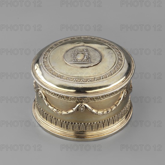 Toilet box, maker: Thomas I Pitts fl. 1755–ca.1793, goldsmith; style: Classicism/Georgian/George III, Classicist; materials: implied metal goldsmith work; date: 1780/1781; place: London, England, Great Britain; form/function: round toilet box with lid toilet accessory; description: decorated with laurel festoons suspended from rosette medallions; lower edge with a knurled belt of stylized lesbian cymatium; slightly arched cover edged with knurled wavy ornament running dog; centrally placed circular medallion with relief of Athena wearing a helmet and holding a spear in her left hand; right side with a circular reserve and an olive branch; ornamentation includes festoons, bay leaves, cymatium, running dog; dimensions: height 5.2 cm, diameter 7.9 cm; weight: 206 g., A round decorative metal box with a domed lid sits on a smooth grey surface; the box appears in shades of gold and silver with a polished reflective finish. The lid features a raised circular medallion with a small sculpted s...