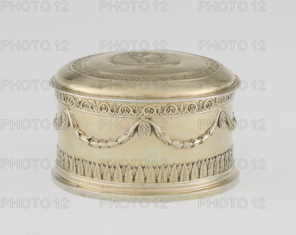 Toilet box; maker: Thomas I Pitts fl. 1755–ca. 1793; goldsmith. Classicism/Georgian, George III style; Classicist toilet box, round with a lid. Decorated with laurel festoons suspended from rosette medallions; lower edge with a knurled belt of stylized Lesbian cymatium. Slightly arched cover edged with a knurled wavy ornament running dog. Centrally placed circular medallion with relief of Athena wearing a helmet and holding a spear in her left hand; on the right side a circular reserve and an olive branch. Ornament: festoons, bay leaves, cymatium, running dog. Inscription/attribution: Ewa Martyna-Michalska. Date: 1780/1781. Place of origin: London, England, Great Britain. Function: toilet accessory. Materials: implied silver/goldsmith work. Dimensions: height 5.2 cm; diameter 7.9 cm; weight 206 g., A small oval metal box with a domed lid, shown against a plain light gray background, rendered in a pale yellowish-gold color with areas of lighter and slightly darker gold tones; the lid ha...