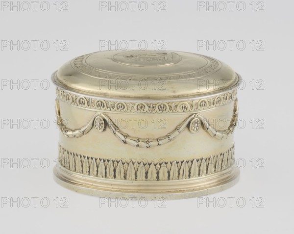 Toilet box by Thomas I Pitts fl. 1755–c.1793, goldsmith; Classicism style; 1780/1781, London, England. Classicist round toilet box with lid, silver, decorated with laurel festoons suspended from rosette medallions. Lower edge has a knurled belt of stylized cymatium; slightly arched cover edged with a knurled wavy running dog ornament. Central circular medallion with a relief of Athena wearing a helmet and holding a spear in her left hand; an olive branch and a circular reserve on the right side. Ornaments include bay leaves and festoons. Toilet accessory; height 5.2 cm, diameter 7.9 cm, weight 206 g., The image shows a round decorative metal box with a lid, primarily in a gold color with subtle variations of pale yellow and light beige; the box has ornate raised garland and floral motifs around its sides and a patterned border near the base and rim.