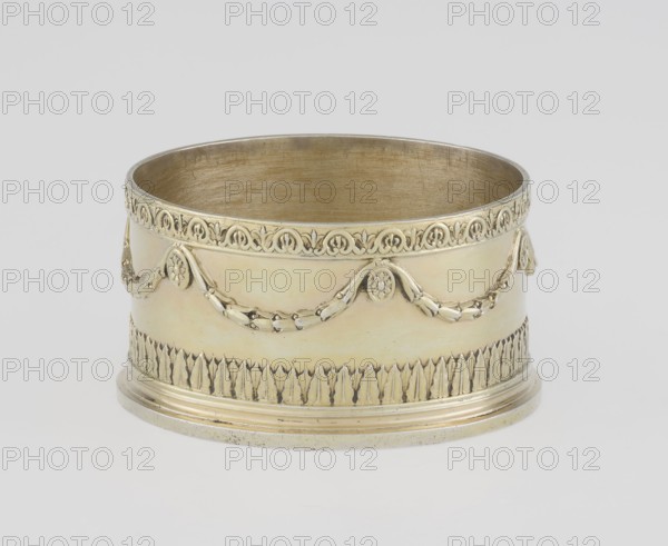 Toilet box, maker: Thomas I Pitts fl. 1755–ca.1793, goldsmith; style: Classicism/George III/Georgian; date: 1780–1781; place: London, England, Great Britain. Classicist round toilet box with lid, decorated with laurel festoons suspended from rosette medallions; lower edge with a knurled belt of stylized lesbian cymatium; slightly arched cover edged with a knurled wavy ornament running dog. Centrally placed circular medallion with relief of Athena wearing a helmet and holding a spear in her left hand; on the right side a circular reserve and an olive branch; additional ornaments: bay leaves, festoons, cymatium. Toilet accessory; height 5.2 cm, diameter 7.9 cm, weight 206 g., A cylindrical metal object with a polished gold tone sits against a light gray background, showing an open top and a rounded base; the exterior features decorative embossed garlands and floral medallions in a matching gold color, a band of repeating ornate motifs near the rim, and a lower frieze of vertical leaf-lik...