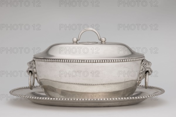 Vase with cover and platter; maker: Marcin Holke fl. ca. 1765–ca. 1792, goldsmith; tableware elements; vases; 18th century; Poland, Warsaw; vessel; height 20 cm, width 46 cm, depth 31.5 cm., A silver lidded serving dish with a rounded lid and arching handle sits on a matching oval tray; the dish and tray have beaded rim decoration, a smooth reflective surface with areas of light and shadow, and sculpted lion-head handles on the sides holding metal rings; visible colors include silver, gray, and subtle reflections of darker gray.