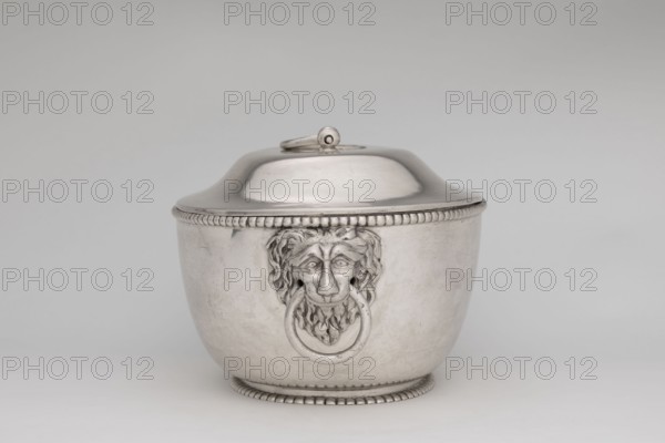 Vase with cover and platter; maker: Marcin Holke fl. ca. 1765–ca. 1792, goldsmith; tableware elements, vases; 18th century; Poland, Warsaw; vessel; height 20 cm, width 46 cm, depth 31.5 cm., A silver-colored metal lidded container with a rounded body and domed lid sits centered against a light gray background, the lid having a small looped handle on top and beaded trim around its lower edge. The body of the container features a decorative lion-head motif in the center holding a round ring in its mouth, and matching beaded trim runs along the base and top rim. The surface of the container is smooth with subtle reflections and faint scuffs, and the overall color palette includes shades of silver, gray, and off-white.