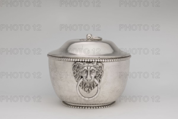 Vase with cover and platter; maker: Marcin Holke fl. ca. 1765–ca. 1792, goldsmith; tableware elements, vases; 18th century; Poland, Warsaw; vessel; height 20 cm, width 46 cm, depth 31.5 cm., A metallic container with a rounded lid and a loop handle on top sits against a plain background, the container showing a raised beaded rim near the lid and a matching beaded base; the front features a sculpted lion head with a large ring held in its mouth, and the surface shows subtle scratches and reflections. The visible colors are silver, gray, and muted off-white in the background.