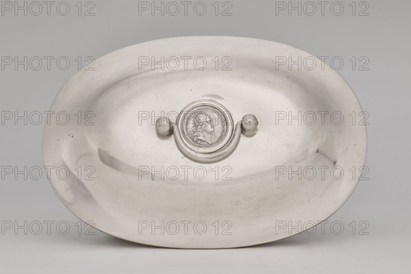 Vase with cover and platter; maker: Marcin Holke fl. ca. 1765–ca. 1792, goldsmith. Tableware elements, vases; 18th century, Poland, Warsaw. Vessel; height 20 cm, width 46 cm, depth 31.5 cm., A shallow oval metal dish in a silver-gray color fills the frame, showing a smooth reflective surface with a few scratches and faint dents; at the center is a raised circular medallion with a sculpted profile of a head in relief, flanked by a curved handle attached to two rounded knobs, all in matching silver-gray tones, against a neutral light-gray background.