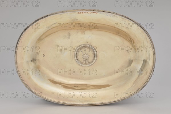 Vase with cover and platter, Marcin Holke fl. ca. 1765–ca. 1792, goldsmith; tableware elements, vases; 18th century; Poland, Warsaw; vessel; height 20 cm, width 46 cm, depth 31.5 cm, An oval shallow dish with a reflective metallic surface showing a warm yellowish-gold tone with areas of paler cream and subtle silvery highlights, a slightly darker golden-brown smudge near the lower rim, and faint grayish tarnish marks and smudges scattered across the interior; a circular raised medallion sits near the center with a central emblem in a slightly darker metallic shade, and the dish has a thin rim with worn darker gray edges.