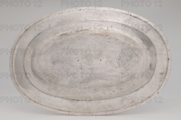 Vase with cover and platter, by Marcin Holke fl. ca. 1765–ca. 1792, goldsmith; tableware elements, vases; 18th century, Poland; Warsaw; vessel; height 20 cm, width 46 cm, depth 31.5 cm, An oval metal tray with a worn, scratched surface in shades of gray and off-white, showing areas of darker gray patina and faint brownish discoloration, a raised rim with a slightly scalloped edge, and a shallow recessed center with visible scuffs and rings.