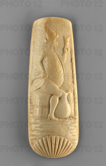Oblong ivory snuff grater lid with rounded corners depicting a man sitting on a barrel, raising a cup in his left hand, his right hand resting on his hip, and a jug at his feet. The grater is topped at the bottom with a semi-plastic shell; the upper end is cut off. 2nd half of the 18th century, Netherlands. Height 13.5 cm, width 4.8 cm., An elongated carved object in a pale cream color with subtle warm beige tones shows a low-relief figure seated on a rounded support, wearing a tall pointed hat and a flowing garment, holding a cuplike vessel in one hand and resting the other hand near the hip; the figure’s legs and detailed shoes are visible above a rounded jug and a barrellike object beside it; the lower portion of the carving features a scallop shell motif with radiating grooves, and the background around the figure has shallow incised texture; the object is photographed against a smooth medium gray backdrop.