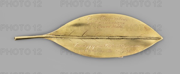 Gold laurel leaf with engraved Polish inscription and date: To President I. Paderewski as a token of homage, the Association of Polish Businessmen in Boltimore, Md., December 1, 1923; maker unknown goldsmith; dedicated to Ignacy Jan Paderewski 1860–1941; 1923; United States of America; height 13 cm, width 4 cm, weight 14 g., A single metallic object shaped like a long leaf rests against a neutral grey background, showing a narrow stem-like handle at one end and a pointed tip at the other; the object has a warm golden color with subtle darker areas and a linear raised midrib running along its length with finely notched lateral veins on either side, and there are a few small surface scratches and slight tarnish variations in the gold tone.