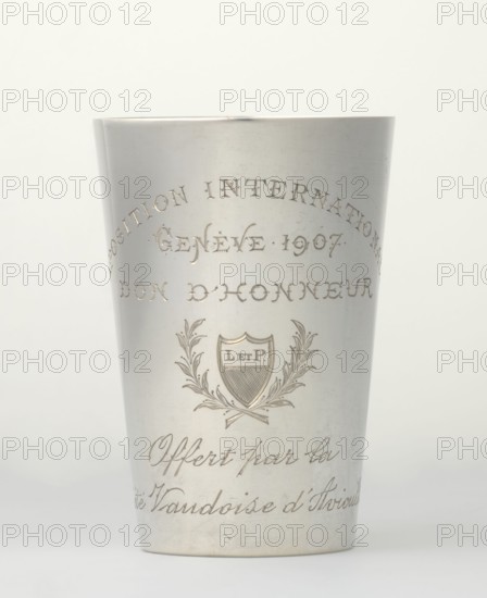 Art Nouveau conical mug by Jezler & Co. Schaffhausen; silversmith; est. 1822. Relief ornament of lily-of-the-valley flowers; engraved inscription: EXPOSITION INTERNATIONALE / GENEVE 1907 / DON D'HONNEUR / cartouche with laurel branches in which the monogram L. ET P. / Offert par la / Societe d'Aviculture. Style: Art Nouveau. Subject: agricultural exhibitions, flowers. Date: 1907, Switzerland; place of origin: Schaffhausen. Materials/technique: silver relief ornament, engraved. Dimensions: height 9 cm, width 6 cm; weight 78 g. Maker: Jezler & Co.; related name: Paderewski, Ignacy Jan 1860–1941., A metal cup with a smooth, slightly tapered cylindrical shape sits against a pale background; the cup has an engraved decorative crest featuring a shield flanked by laurel-like branches and a ribbon element, with several lines of engraved script above and below the crest. The surface of the cup is metallic silver with subtle reflections and areas of light patina or wear, and the background is of...