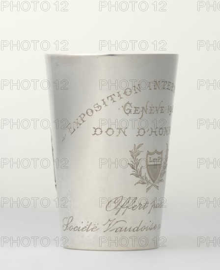 Art Nouveau conical mug by Jezler & Co Schaffhausen; silversmith; est. 1822. Relief ornament of lily of the valley flowers; engraved inscription: EXPOSITION INTERNATIONALE / GENEVE 1907 / DON D'HONNEUR and cartouche with laurel branches containing monogram L. ET P. and italic Offert par la / Societe d'Aviculture. Dated 1907, Switzerland Schaffhausen. Height 9 cm, width 6 cm, weight 78 g., A cylindrical metallic cup with a smooth light gray surface stands upright against a pale off-white background, showing engraved decorative elements including a laurel-like emblem and flowing script-like ornamentation; the metal has subtle wear marks and faint scratches, and the overall tones visible are silver-gray, light gray, off-white, and a hint of darker gray in the engraved lines.