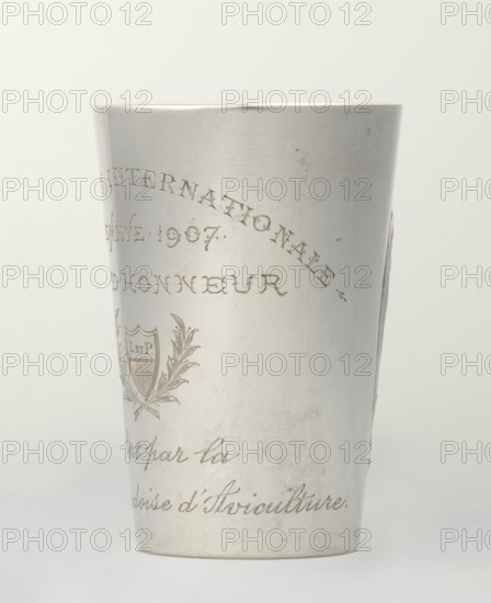 Art Nouveau conical mug with relief ornament of lily-of-the-valley flowers and engraved inscription EXPOSITION INTERNATIONALE / GENEVE 1907 / DON D'HONNEUR with a cartouche of laurel branches containing the monogram L. ET P. and the italic text Offert par la / Societe d'Aviculture. Maker: Jezler & Co. Schaffhausen; silversmith; est. 1822. Associated name: Paderewski, Ignacy Jan 1860–1941. Style: Art Nouveau. Event/date: Exposition Internationale, Geneva, 1907. Materials/measurements: silver; height 9 cm, width 6 cm, weight 78 g. Origin: Schaffhausen, Switzerland., A tall cylindrical metal cup with a smooth, slightly reflective surface and a matte silver color, shown against a pale off-white background; the cup displays a decorative engraved crest and flowing engraved script around its body, with small areas of light scuffs and subtle scratches visible on the metal, and faint variations in tone ranging from bright silver highlights to slightly darker silvery-gray areas, while the interi...
