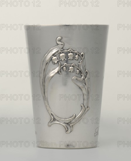Art Nouveau conical mug by Jezler & Co Schaffhausen; silversmith; established 1822. Relief ornament of lily-of-the-valley flowers; engraved inscription EXPOSITION INTERNATIONALE / GENEVE 1907 / DON D'HONNEUR and a cartouche with laurel branches containing the monogram L. ET P. plus italic Offert par la / Societe d'Aviculture. Dated 1907, Switzerland Schaffhausen., Art Nouveau. Dimensions: height 9 cm, width 6 cm; weight 78 g., A cylindrical metallic cup with a smooth, reflective silver surface stands against a pale off-white background, featuring an ornate raised decorative motif on its front that resembles flowing, curved leaves and bell-shaped flowers in the same silver tone, with the decoration forming an open oval frame and curling flourishes; subtle scratches and small tarnished spots appear on the metal, and the overall color palette includes shades of silver, light gray, and the neutral off-white of the background.