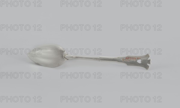 Dessert spoon, Jarra, Marcin 1852–1938 goldsmith; Dessert spoon with an ovoid, smooth, pointed scoop, a stepped handle ending in a stylized crown and decorated with an engraved geometric ornament stylized to resemble Zakopane style; early 20th century; Poland, Kraków; Zakopane style; cutlery; height 16 cm, width 3 cm, weight 35 g., A metallic spoon lies horizontally on a light gray background, showing a slightly rounded bowl with a smooth reflective surface and a long flat handle that ends in a decorative flattened tip with cutout shapes; the spoon appears silver in color with slight darker gray shadows and subtle highlights, and there is a small area of reddish discoloration near the handle tip.