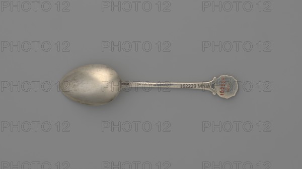 Souvenir spoon, Ford Company fl. 1875–1899. Souvenir spoon with an ovoid scoop with a pointed tip; half-handle with oblique notches, then flat, splitting at the end with a mounted circular plaque with a struck facsimile of the seal of Yale University — in the center an open book with the Hebrew inscription URIM./TUMMIM enclosed in a cartouche around which is the Latin inscription LUX ET VERITAS; inscription around the rim: SIGILL: COLL: YALEN: NOV: PORT: NOV: ANGL:. 1890s. New Haven, United States of America. Cutlery. Height 10.5 cm; width 2.4 cm; weight 15 g., A single metal spoon lies flat on a plain gray background, showing a rounded bowl with a slightly worn and mottled silver surface and a long, slim handle ending in a scalloped decorative tip; visible colors include various shades of silver, gray, and a small area of muted red on the decorative tip.