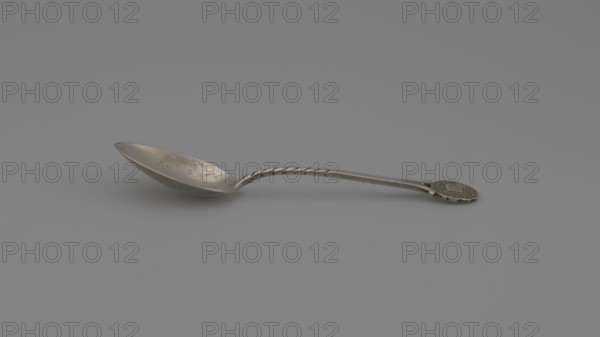 Souvenir spoon, Ford Company fl. 1875–1899 role classification of creator, goldsmith's company author, Paderewski, Ignacy Jan 1860–1941 — Souvenir spoon with an ovoid scoop with a pointed tip; half-handle with oblique notches, then flat, splitting at the end with a mounted circular plaque with a struck facsimile of the seal of Yale University in New Haven — in the center an open book with the Hebrew inscription URIM./TUMMIM enclosed in a cartouche around which is the Latin inscription LUX ET VERITAS motto; inscription around the rim: SIGILL: COLL: YALEN: NOV: PORT: NOV: ANGL:, 1890s, United States of America, New Haven, cutlery, height 10.5 cm, width 2.4 cm, weight 15 g, A metal spoon lying on a smooth gray surface, with a long twisted handle and a shallow bowl showing some mottled darker spots; the spoon and surface are shades of silver and gray, with subtle darker gray shadows beneath the spoon and a hint of brownish tarnish on the bowl.