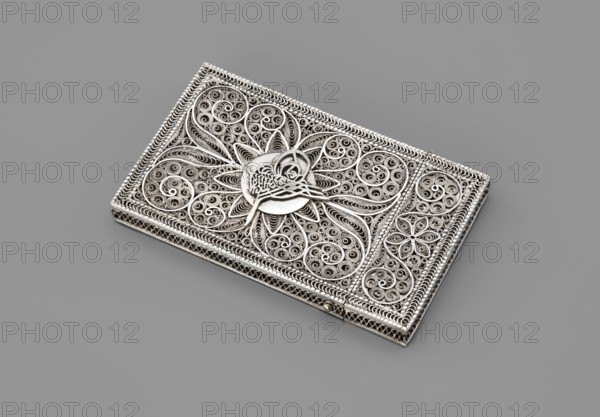 Business card holder, unknown goldsmith; Paderewski, Ignacy Jan 1860–1941 mentioned in provenance; inscriptions in Kufic script. Business card holder in the form of a flat rectangular box, silver filigree with plant motifs and Kufic inscription. Late 19th century, possibly Germany. Smoking accessories/varia. Dimensions: height 10 cm, width 6 cm, depth 1 cm; weight 80 g., A rectangular ornate metallic object lies on a flat surface; it has intricate filigree work with swirling and looping patterns, floral and petal-like motifs, and a raised central medallion featuring a stylized bird figure. The piece appears silver with darker shadowed recesses that emphasize the detailed texture, and the background surface is a uniform gray.