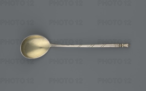 Decorative spoon, Dmitriyev, M fl. 1854–1877 goldsmith; Decorative spoon with a teardrop-shaped scoop, decorated on the reverse with a polished niello acanthus twig; angular handle at the base, becoming conical, ending with a bowl. 1863, Moscow, Russia; cutlery; height 13 cm, width 2.8 cm, weight 20 g., A single metal spoon lies horizontally against a solid muted gray background, showing a rounded bowl with a warm golden interior and a cooler silvery outer rim; the long narrow handle is silver with subtle linear grooves and a twisted decorative band near the end, which terminates in a small turned finial that is slightly darker.