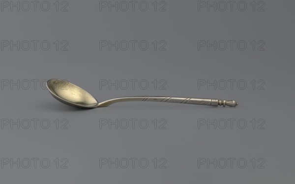Decorative spoon by Dmitriyev, M fl. 1854–1877, goldsmith. Decorative spoon with a teardrop-shaped scoop, decorated on the reverse with a polished niello acanthus twig; angular handle at the base, becoming conical, ending with a bowl. Possibly dated 1863; Moscow, Russia. Materials/technique: niello/cutlery. Dimensions: height 13 cm, width 2.8 cm, weight 20 g., A single metal spoon rests on a flat surface against a gray background, showing a shallow, oval bowl with a slightly worn, yellowish metallic interior and a darker silver exterior; the long handle is slender and silver with subtle diagonal grooves and a small rounded finial at the end in a slightly darker silver tone, and there is a faint shadow beneath the spoon.