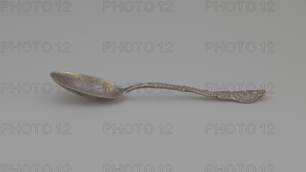 Souvenir spoon, Gorham Mfg. Co. Providence; goldsmith's manufactory; fl. 1865-, Paderewski, Ignacy Jan 1860-1941 — Eads, James Buchanan 1820-1887, portraits, New Orleans United States of America, landscapes. Souvenir spoon with an oval scoop with a relief representation of harbor fortifications, bay and ships; at the edge of the scoop the inscription EADS JETTIES. NEW ORLEANS; handle with an irregular edge bordered with acanthus leaves; in the finial a portrait of a man with a beard and at the bottom the signature on the sash CAPT. EADS, below the breakwater; late 19th century; Providence, United States of America; cutlery; height 14.8 cm, width 3 cm, weight 30 g., A single metal spoon rests on a smooth, light gray surface against a slightly darker gray background; the spoon has a rounded bowl with a subtle patina showing darker and lighter areas, and a long ornately detailed handle with raised decorative patterns and a slightly darker metallic tone overall, with visible highlights and...