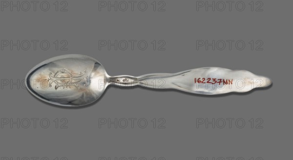 Souvenir spoon, Whiting Manufacturing Co. fl. 1866–1926. Souvenir spoon with egg-shaped scoop featuring a representation of the Capitol building in Washington; flat handle with a relief lily of the valley sprig; engraved IP monogram tied on the reverse. Lily of the Valley pattern introduced in 1885. Late 19th century. Bridgeport, United States of America. Cutlery; height 15 cm, width 3.1 cm, weight 25 g. Maker role: goldsmith/company., A single metal spoon lies horizontally against a solid gray background, showing a reflective bowl with an engraved decorative motif and some darker tarnish areas, a narrow neck with a slight twist, and a gently scalloped handle that has smoother and slightly worn patches; visible colors include silver tones, darker gray reflections, warm brownish tarnish, and the neutral gray of the background.