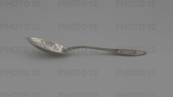 Souvenir spoon, Towle Silversmith fl. 1882-, role classification of maker, goldsmith company author, Paderewski, Ignacy Jan 1860-1941 — souvenir spoon with an ovoid dipper with a pointed tip, with a scene depicting the interior of a mine with two men with pickaxes; on the reverse an engraved inscription in cursive Batle Mont probably referring to Battle Mount, a city in Nevada; straight, flaring handle with a lanceolate tip, handle grip with a depiction of a man standing by a well, turning a crank, winding up a chain running along the handle, to which a bucket filled with ore is attached at the end, reaching into the mine; on the reverse of the handle a crossed shovel, pickaxe and a rope with an incomprehensible inscription; late 19th century; United States of America; cutlery; height 14.6 cm, width 2.3 cm, weight 30 g, A single metal spoon lies horizontally on a flat surface, showing a reflective bowl with decorative embossed patterns and a gently curved handle with a beaded edge and ...