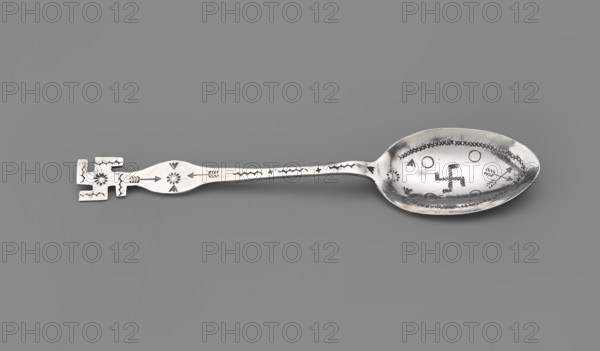 Souvenir spoon, subject: Paderewski, Ignacy Jan 1860–1941 inscription or association. Souvenir spoon with an ovoid scoop, flat handle widening at the end and finished with a cut-out swastika; scoop decorated with engraved motifs of circles, sun, crossed arrows and a swastika in the middle, similar motifs on the handle. Probably made on one of the Indian reservations possibly Navajo, late 19th century, United States of America. Cutlery; height 14 cm, width 3 cm, weight 10 g., A silver-colored metal spoon lies horizontally against a solid gray background, with an ornate squared handle on the left featuring angular cutouts and stamped decorative motifs, a slightly tapered and engraved stem with arrow and floral-like stamped designs, and an oval bowl on the right that shows a patterned stamped border and several stamped circular and linear markings; visible colors include silver and shades of gray in the background.