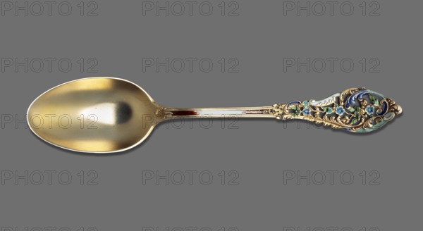 Tea spoon, Dominick & Haff fl. 1868–1928. Trianon pierced cutlery model, Neo-Rococo style. Tea spoon with an ovoid, smooth scoop with a knotted HP monogram engraved on the reverse; straight, flaring, openwork handle with Neo-Rococo ornament featuring rocailles, acanthus and small flowers; interior of handle enamelled in various shades of white, green, blue; brown enamel along the handle fading to white. The Trianon pierced handle pattern was introduced by the company in 1885; the HP Helena Paderewska monogram indicates the spoon was purchased or given no earlier than 1899 the Paderewskis' wedding date. Maker active in New York, United States. Cutlery. Dimensions: height 15.5 cm, width 3.1 cm, weight 20 g., A single ornate spoon lies against a solid gray background, with a large rounded bowl that has a warm golden hue and subtle reflective highlights, and a long handle that transitions from a polished metallic shaft into an intricately decorated end featuring swirling gold-toned filigre...