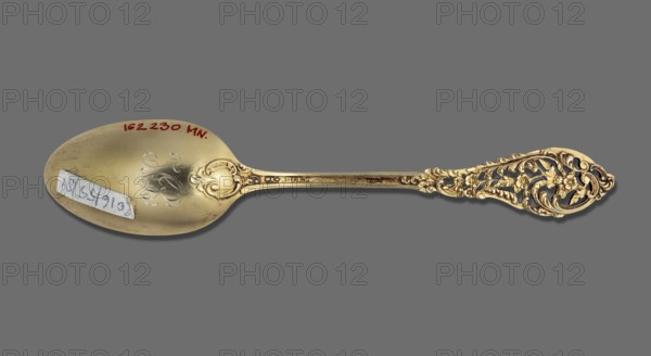 Tea spoon, Dominick & Haff fl. 1868–1928. Trianon pierced cutlery model, Neo-Rococo style. Tea spoon with an ovoid, smooth scoop with a knotted HP monogram engraved on the reverse; straight, flaring, openwork handle with Neo-Rococo ornament of rocailles, acanthus and small flowers, enamelled on the inside with colored enamel in shades of white, green, blue; brown enamel along the handle, fading to white. The Trianon pierced handle pattern was introduced by the company in 1885; the HP Helena Paderewska monogram indicates the spoon was purchased or given no earlier than 1899 the Paderewskis' wedding date. New York, United States of America. Cutlery. Height 15.5 cm; width 3.1 cm; weight 20 g., A decorative metal spoon lies horizontally against a solid gray background, showing a warm golden surface with areas of darker tarnish and lighter highlights; the bowl of the spoon has a subtle engraved swirl pattern and a small rectangular paper label affixed, and the handle features ornate openwor...