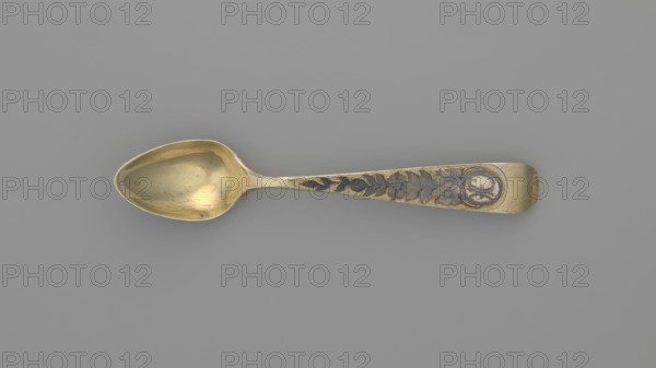 Tea spoon by Fedor Klimov Buszkowskij fl. 1795–1834, goldsmith. Decorative gilded spoon with an ovoid scoop with a pointed tip; reverse decorated with a niello ornament of fish scales and rocailles. Flat handle widening at the end, slightly bent and rounded, with niello decoration on both sides with floral motifs. Incised coats of arms. Ornament: flowers, fish scale. Date: 1815. Place of origin: Veliky Ustyug, Russia. Material/function: cutlery tea, teaspoon. Dimensions: length 14 cm, width 2.6 cm, weight 15 g., A single tablespoon with a rounded bowl and a tapered handle lies flat against a plain background; the spoon has a metallic sheen in gold and brass tones with darker grayish areas in the recessed decorative elements, and the handle features a floral and leaf pattern in contrasting darker gray set into the gold surface, ending in a rounded tip with a circular recessed area; the background is a uniform medium gray.
