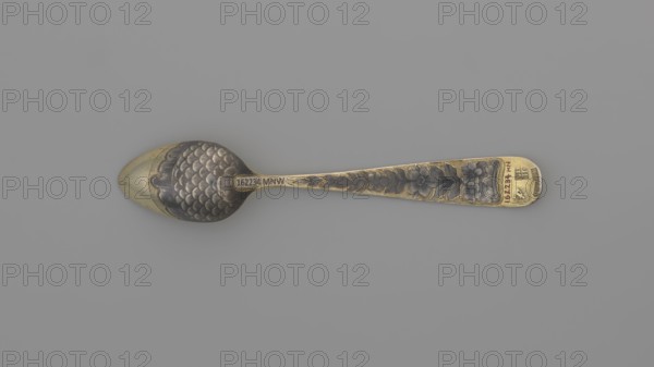 Tea spoon by Buszkowskij, Fedor Klimov fl. 1795–1834, goldsmith; decorative gilded spoon with an ovoid scoop with a pointed tip, reverse decorated with a niello ornament of fish scales and rocailles; flat handle widening at the end, slightly bent and rounded, with niello decoration on both sides with floral motifs. Inscribed/date: 1815. Place of origin: Veliky Ustyug, Russia. Subject/ornament: flowers, fish scale pattern, coats of arms. Function/materials: tea cutlery, tea spoon; length 14 cm, width 2.6 cm, weight 15 g., A single ornate spoon lies horizontally on a plain gray background, showing a metallic surface with gold and darker silver tones; the bowl of the spoon has a scalloped, scale-like textured pattern in dark silver with a gold rim and a smooth gold tip, while the handle tapers to a rounded end and features raised floral and leaf decorations in dark silver against a gold base, with the decorated areas appearing slightly darker and aged.