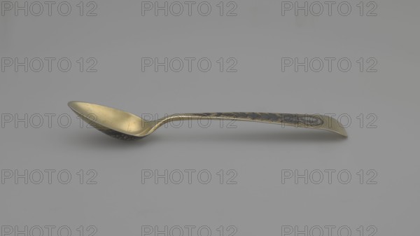 Tea spoon by Buszkowskij, Fedor Klimov fl. 1795–1834, goldsmith. Decorative gilded spoon with an ovoid scoop with a pointed tip; reverse decorated with a niello ornament with fish scales and rocailles. Flat handle, widening at the end, slightly bent and rounded, with niello decoration on both sides with floral motifs. Ornament: flowers, fish scale; coats of arms. Tea cutlery. Date: 1815. Place of origin: Veliky Ustyug, Russia. Materials/function: cutlery; length 14 cm, width 2.6 cm, weight 15 g., A single spoon lies horizontally on a plain background, showing a curved bowl and a slightly upturned handle with decorative patterns; the spoon's surfaces display metallic colors including gold and dark gray, and the background appears light gray.