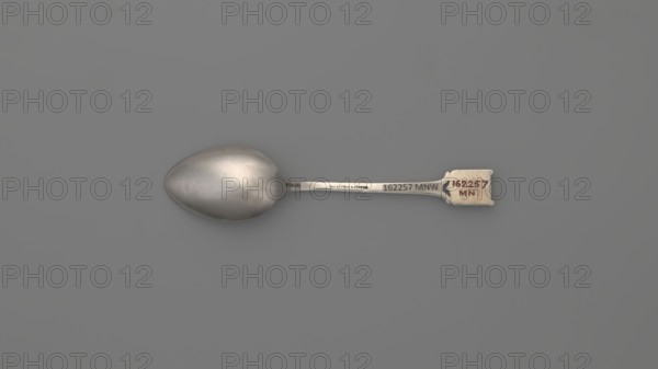 Souvenir spoon, Ford Company fl. 1875–1899. Souvenir spoon with an ovoid scoop with a pointed tip; half-handle with oblique cuts, then flat, splitting at the end, with a plaque attached in the form of a cartouche with an open book with the Hebrew inscription URIM/TUMMIM on a background of sapphire enamel; corporate slogans/goldsmith company attribution; maker associated with Paderewski, Ignacy Jan 1860–1941 noted. 1890s, New Haven, United States of America. Cutlery. Length 10.5 cm; width 2.4 cm; weight 15 g., A single metal spoon lies horizontally against a flat gray background, showing a smooth, slightly reflective bowl on the left and a straight handle extending to the right that ends in a small rectangular tab; the spoon surfaces display shades of silver and gray with subtle highlights and darker silver tones, and there are small areas of light beige and muted reddish-brown on the tab at the handle end.