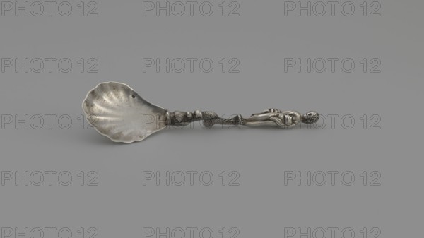 Decorative silver spoon by François-Auguste Boyer-Callot fl. 1865-, 4th quarter of the 19th century, Paris, France. Silver spoon with a shell-shaped scoop and a handle formed as a solid dolphin supporting a naked woman Venus who covers her breasts with her left hand and holds a shawl to cover her womb in her right. Motif includes dolphins symbol of Venus. Cutlery; varia. Length 8.5 cm; width 2.4 cm; weight 10 g., The image shows a single ornate spoon lying horizontally on a flat background. The spoon has a scallop-shaped bowl with a slightly worn, metallic surface and visible surface texture. The handle is elaborate, featuring a sculpted figure at the end and a segmented, decorative shaft with textured details. The visible colors are shades of gray and silver.