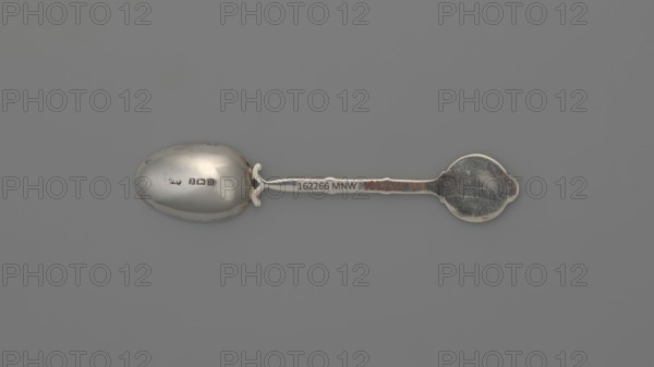Decorative spoon by Deakin & Francis Ltd Birmingham; goldsmiths; fl. from 1786. Spoon with an ovoid scoop, smooth; handle formed of two intertwined stylized branches with leaves, ending in a round flat terminal with enameled decoration: on a white background a branch with yellow spherical fruits and lanceolate green leaves. Cutlery; height 11.5 cm, width 2.4 cm, weight 15 g; dated 1912/1913; England, Birmingham, Great Britain., A single metal spoon lies horizontally against a flat gray background, showing a smooth oval bowl on the left and a rounded, slightly scalloped handle end on the right; the spoon has a worn, reflective silver surface with visible scratches and patches of darker tarnish, and there are small areas of duller and shinier silver along the stem and bowl.