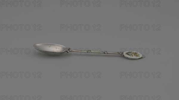Decorative spoon, Deakin & Francis Ltd Birmingham; goldsmith's firm; fl. 1786- — paderewski, Ignacy Jan 1860–1941 — decorative spoon with an ovoid scoop, smooth; handle formed of two intertwined stylized branches with leaves, ending in a round flat terminal with enameled decoration: on a white background a branch with yellow spherical fruits and lanceolate green leaves. 1912/1913, Birmingham, Great Britain. Cutlery; height 11.5 cm, width 2.4 cm, weight 15 g., A metal spoon with a patinated silver-colored bowl and a long slender handle lies on a smooth gray surface; the handle has a textured, segmented design and ends in a round decorative disc that shows a floral motif with green leaves and yellow blossoms on a white background.