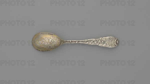 Egg spoon, Tiffany & Co. New York; goldsmith and dealer; founded 1853. Maker/designer: Charles Grosjean 1841–1888. Chrysanthemum pattern, introduced by the company in 1880. Late 19th century. Egg spoon with an ovoid scoop, gilded inside, with a profiled, widening handle topped with a chrysanthemum flower; both sides of the handle bear a relief decoration of chrysanthemum leaves and flowers, overlapping the scoop on the back. Materials: cutlery. Dimensions: length 11.5 cm; width 2.5 cm; weight 30 g., A single ornate spoon lies horizontally against a smooth gray background, showing a metallic surface with a warm golden tone on the bowl and a cooler silver tone along the handle; the spoon features intricate raised floral and leafy relief work extending from the bowl onto the handle, with flowing curved lines and petal-like shapes, and a slightly darker patina in recessed areas that accentuates the decorative details.