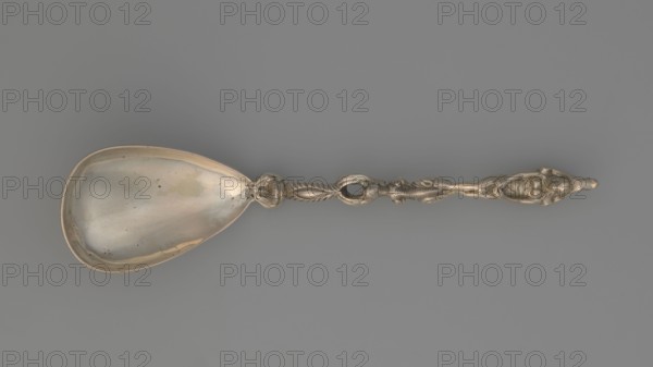 Decorative spoon, Decorative spoon with a teardrop-shaped, smooth scoop and a handle decorated with a mascaron at the base, topped with a figure of a naked woman standing on a snake, shielding her womb with her hands and a stylised leaf; on the back of the ladle engraved inscription L. ALMA TADEMA Lawrence or his daughter Laurence, end of the 19th century, Germany, cutlery, varia; height 18.5 cm, width 4.5 cm, weight 75 g., A long ornate spoon lies horizontally against a flat gray background; the spoon has a shallow, teardrop-shaped bowl with a reflective, slightly tarnished surface showing subtle warm highlights and pale greenish spots, and an intricately worked handle featuring twisted rope-like elements, sculpted floral and foliate motifs, and a carved figure at the terminal. The visible colors are metallic silver with warm golden reflections, pale greenish discoloration inside the bowl, and the neutral gray of the background.