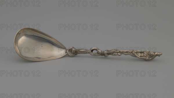 Decorative spoon with a teardrop-shaped, smooth scoop and a handle decorated with a mascaron at the base, topped with a figure of a naked woman standing on a snake, shielding her womb with her hands and a stylised leaf; on the back of the ladle engraved inscription L. ALMA TADEMA Lawrence or his daughter Laurence, end of the 19th century, Germany, cutlery, varia; height 18.5 cm, width 4.5 cm, weight 75 g, A metal spoon lies horizontally against a smooth gray background, with a wide, shallow bowl on the left showing reflective surfaces and subtle blemishes, and a decorative handle on the right featuring an ornate, sculpted design with twisted and rounded elements; visible colors include silver tones, gray, and faint hints of warm metallic reflections.