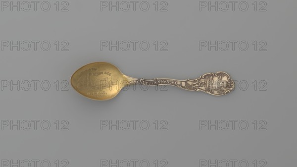 Souvenir spoon, Shepard Mfg. Co. fl. 1893–1923. Silver souvenir spoon, ovoid scoop with engraved view of buildings and the inscription JACKSONS / BIRTHPLACE / NASHVILLE / TENN. Flat handle with irregular edges made of channel sections; the inscription TENNESSEE along the handle, above which is a man with a pickaxe. The handle is widened at the end with relief depictions of a plow, a sheaf of wheat above it the Roman numeral XVI, cotton, with the inscription AGRICULTURE underneath, and a ship below with the inscription COMMERCE below — the emblems of the state of Tennessee; XVI denotes the state's serial number; AGRICULTURE and COMMERCE are the state motto. Depicts Paderewski, Ignacy Jan 1860–1941 and references Andrew Jackson 1767–1845. 19th/20th century. Melrose Highlands, United States of America. Cutlery. Length 9.5 cm; width 2.2 cm; weight 10 g., A decorative metal spoon rests on a flat surface, showing a bowl with a warm golden color and a handle with darker metallic tones; the ha...