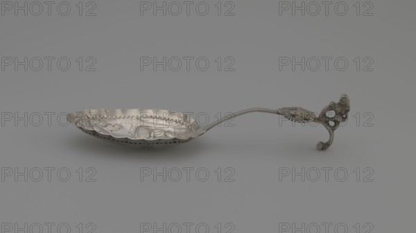 Decorative spoon, oval scoop with a relief pastoral scene framed in a pearled border — a woman sits under a tree with two sheep beside her; floral decoration at the bottom; irregular, wavy scoop edge; flat handle with a volute ending, decorated with an openwork section depicting a winged putto head and a sculpted lion standing on its hind legs; late 19th century, Netherlands; cutlery; height 14 cm, width 4.3 cm, weight 20 g., A metallic spoon-like object rests on a gray surface, showing a shallow, oval bowl with a scalloped rim and decorative embossed patterns inside; the bowl has a row of small rounded indentations near its edge and a series of small perforations along its lower edge. A slender, gently curved handle extends from the bowl to an ornate end piece with a curled, openwork design and textured details. The visible colors are shades of silver and gray, against a muted gray background.