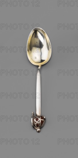 Souvenir spoon by Fenton, James Birmingham; goldsmith's firm; fl. 1852–1954. Spoon with an ovoid scoop gilded inside and an angular handle ending with an openwork sword hilt set with an agate. Late 19th century, Birmingham, England. Cutlery; height 10.5 cm, width 2.8 cm, weight 10 g. Maker attribution includes Paderewski, Ignacy Jan 1860–1941 noted in inscriptions/decorative references., A single spoon is shown against a gray background, with a reflective bowl that appears silver with golden tones inside, a long silver-colored handle, and an ornate end featuring a small spherical element and a decorative cage-like piece with reddish-brown visible through the openwork; the spoon shows highlights and reflections of darker shapes on its surface.