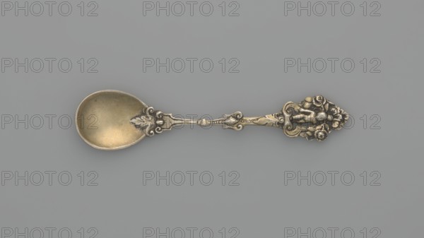 Decorative spoon, Neresheimer Hanau; goldsmith's workshop; 1890-, goldsmith's company author, Paderewski, Ignacy Jan 1860-1941 - testament provenance, grotesques, neo-Renaissance style, putti. Decorative spoon with a circular scoop and a profiled handle topped with a grotesque with a putti standing in an arcade decorated with roses, late 19th century, Germany, Hanau, cutlery, varia; length 11 cm, width 2.4 cm, weight 20 g, A single ornate metal spoon lies horizontally on a flat background, showing a bowl with a slightly worn golden tone and a decorative handle with intricate raised floral and scroll motifs in a darker silvery and warm brass color; the background is a smooth muted gray.