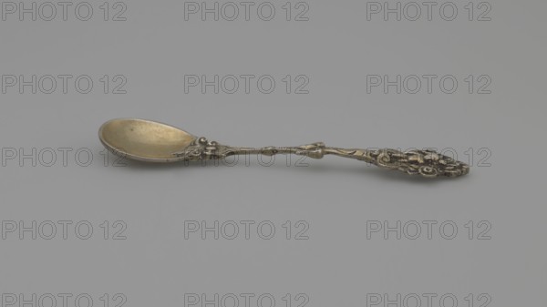 Decorative spoon, Neresheimer Hanau; goldsmith's workshop; 1890-, goldsmith's company; grotesques, neo-Renaissance; putti. Decorative spoon with a circular scoop and a profiled handle topped with a grotesque with a putti standing in an arcade decorated with roses, late 19th century, Germany, Hanau, cutlery, length 11 cm, width 2.4 cm, weight 20 g., A single ornate spoon lies on a smooth background, showing a shallow bowl with a warm golden tone and a handle with intricate raised decorative patterns in a darker metallic shade; the overall colors visible are gold, silver, gray, and subtle darker accents, with the background appearing as a uniform light gray.