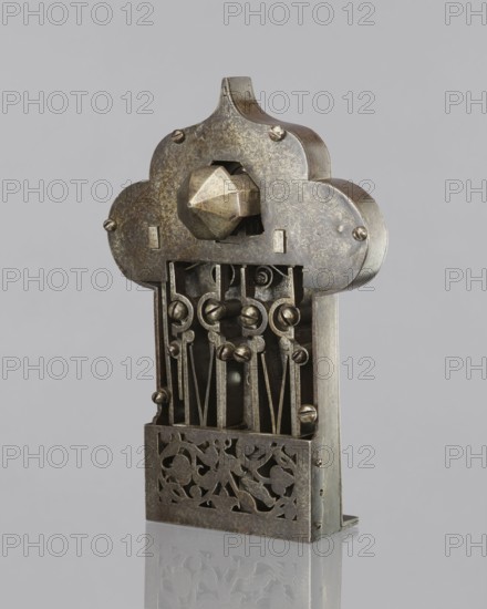 Four-bolt door lock with key. A lock of fine locksmithing workmanship, probably 17th century, Baroque style. Rectangular plate with a trefoil rear section; cover engraved with a twig motif. Side bulges with profiled male busts with mustaches and a central en face mask with a hole for the register dome. Plate bent at a right angle for mounting on the door edge; mechanism partially covered by an openwork cover engraved with a twig motif featuring a bird and a pomegranate. Screw mounting. The key is missing and the brass box cover that formerly covered the central part of the lock is absent. Metalwork; height 33 cm, width 22 cm, depth 12.8 cm., An ornate metal mechanism stands against a pale gray background, showing a weathered metallic surface with tones of gray, silver, and hints of brownish patina; the top has a rounded, cloudlike silhouette with a faceted geometric knob projecting from its center and small rounded fasteners at several points; below, a visible interior of vertical rods...