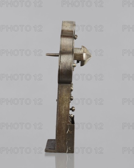 Four-bolt door lock with key missing. 17th century. Baroque style; metalwork. Rectangular plate with a trefoil rear section; cover engraved with a twig motif. Side bulges bear profiled male busts with mustaches and a central en face mask with a hole for the register dome. Plate is bent at a right angle for mounting on the door side; mechanism partially covered by an openwork cover engraved with a twig motif featuring a bird and a pomegranate. Screw mounting. Brass box cover that once covered the central part is missing. Height 33 cm, width 22 cm, depth 12.8 cm. High-quality locksmithing workmanship with precise finishing; the key and its richly decorated register was originally meticulously crafted., A vertical metal object photographed against a light gray background, shown in profile; the object is mostly a dull silvery-gray with areas of darker gray and brownish tarnish, featuring a rounded top section with a pointed arrow-like protrusion and a short horizontal rod, a flat base with...