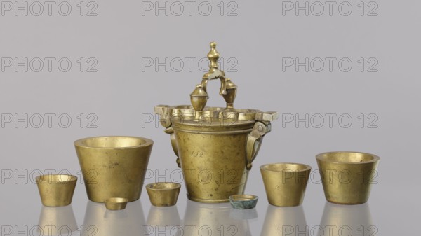 Set of weights, Seven weights in the form of bowls—truncated cones—and an eighth, largest piece serving as a container for the others, with a hinged lid closed by a hasp and a movable handle mounted in pinnacles shaped like simplified vases. On the lid: 16 MARK and a Prussian eagle; hallmark BERLIN; another mark engraved 160; 152 engraved on the side weight 1865.57 g. 18th century, Germany. Weight; height 17.5 cm, width 10.2 cm, depth 14.3 cm., A group of metal vessels arranged on a reflective surface with a plain light gray background, including a central ornate vessel with a decorative handle and two small finials, flanked by several simpler cup-shaped vessels of varying sizes and shapes. The objects show a metallic gold color with areas of darker patina and small spots of greenish corrosion on one smaller piece, and faint reflections of the items appear on the surface beneath them.